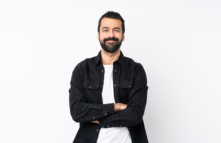 A man with beard and black shirt standing in front of white wall.